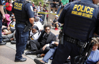 Cornel West, center, joins other protesters sitting on the steps of the Thomas F. Eagleton Federal Courthouse as members of the Federal Protective Service stand watch Monday, Aug. 10, 2015, in St. Louis. Protesters have been arrested after blocking the entrance to a St. Louis federal courthouse while calling for more aggressive U.S. government response to what they call racist law enforcement practices. (AP Photo/Jeff Roberson)